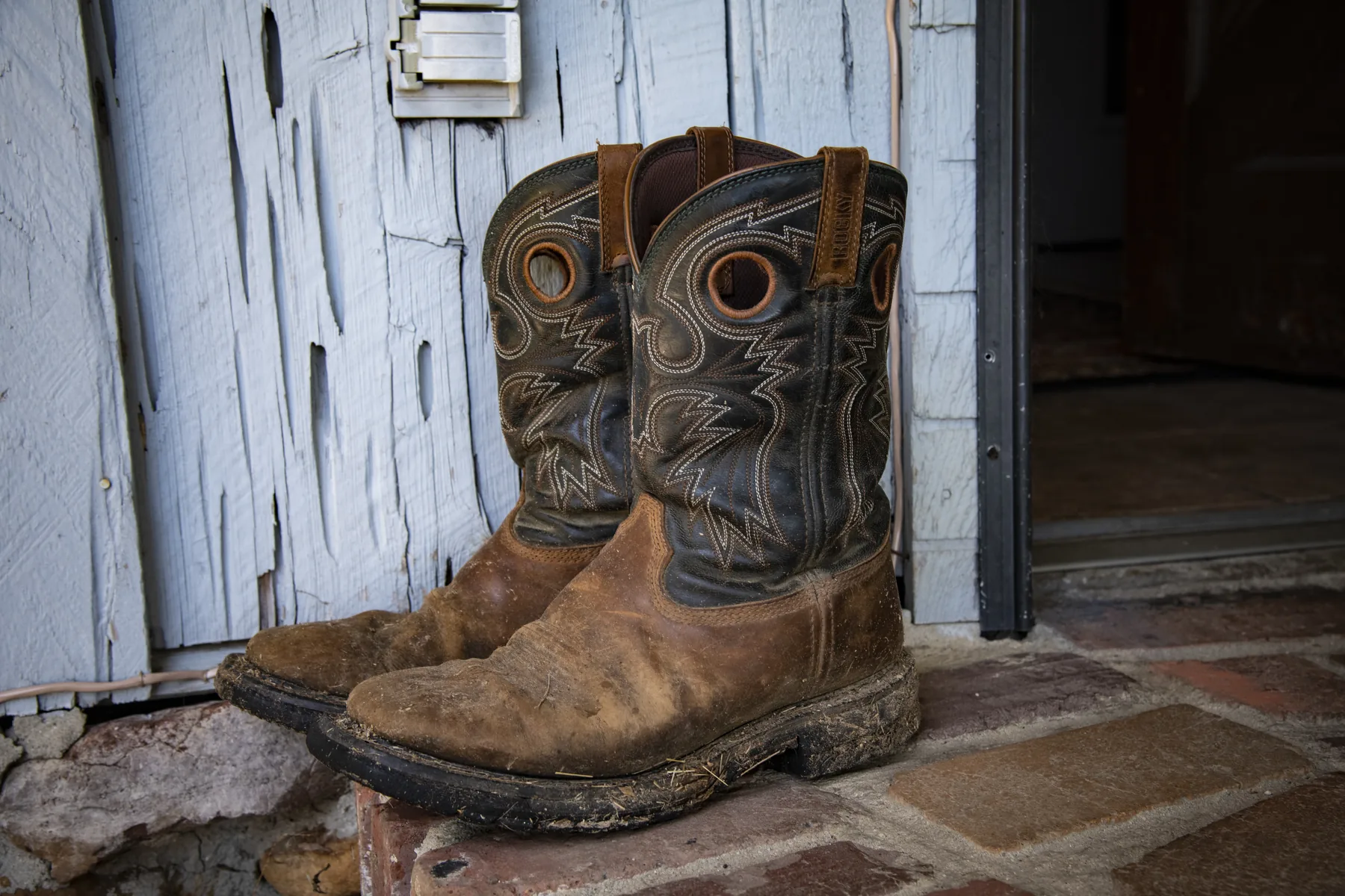 A worn pair of work boots left standing beside an open barn
                  door, white paint peeling off the boards.